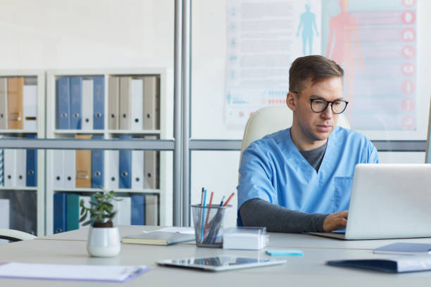 A man in blue scrubs sitting at a desk with a laptop.